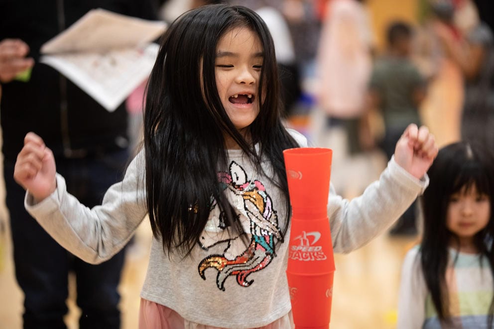 A child smiles and celebrates behind a tall tower of cups stacked end-on-end.