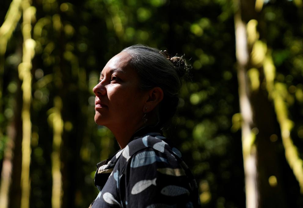A person poses for a portrait and looks off camera in front of a dense curtain of trees.