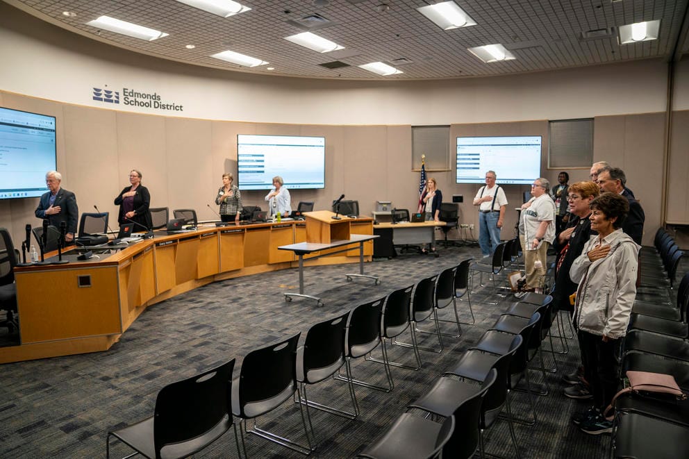 Board members, staff, and members of the public recite the Pledge of Allegiance during an Edmonds School Board meeting.