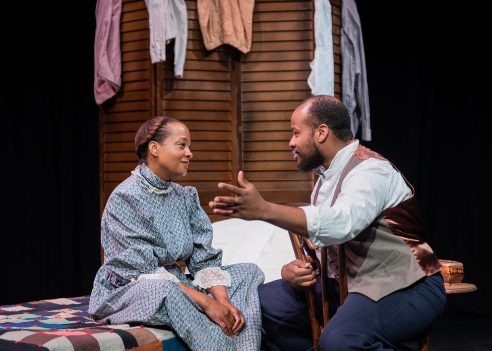 A man and woman sit on a bed and talk to each other. They're both wearing 19th century ranch clothing.