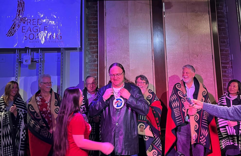 A man on stage puts on a Denver Broncos necklace while a crowd of people smile behind him