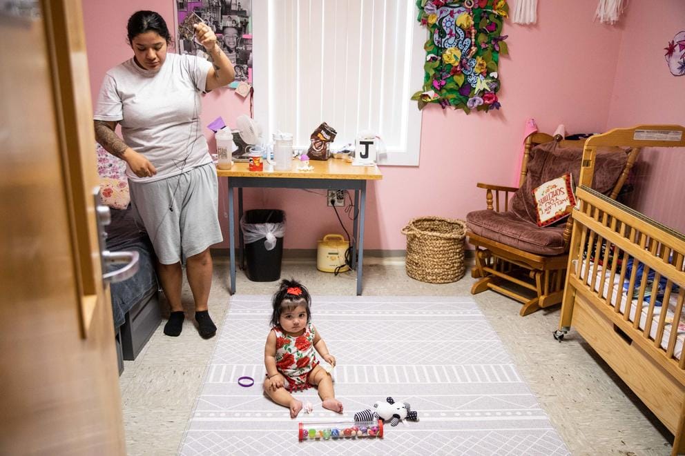 A baby looks up from the floor of a pink bedroom with a bed and a crib. Christina Torres stands to the left.