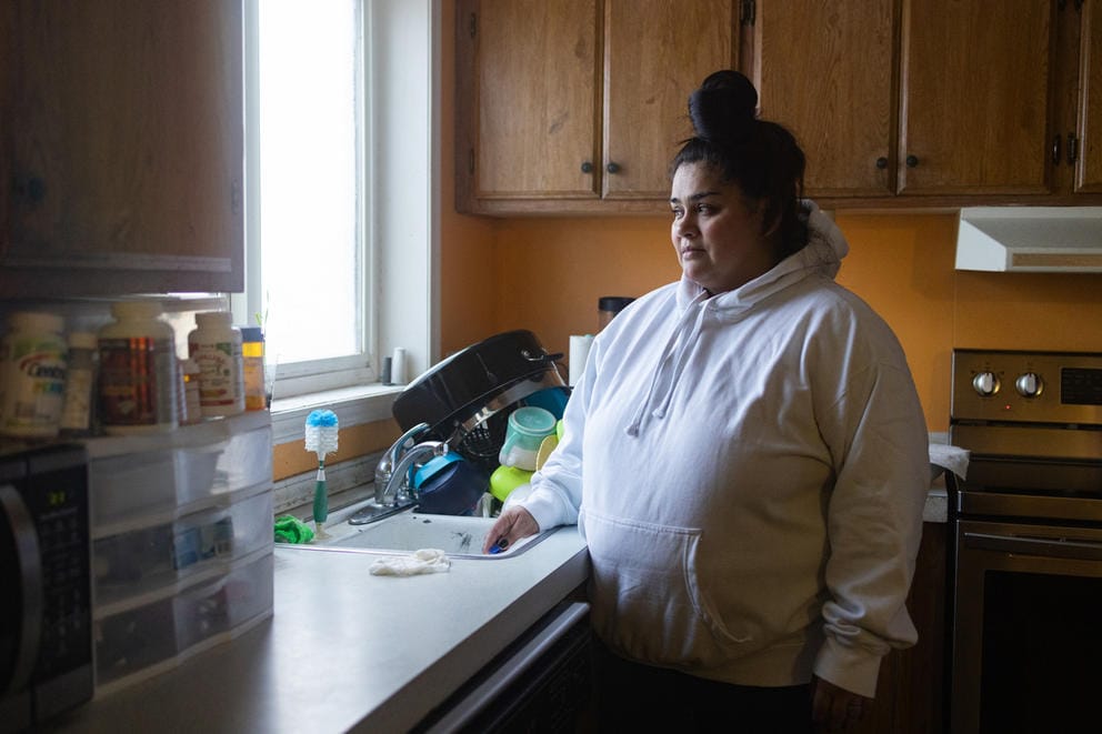 A woman looks out the window of her mobile home in Moxee, Washington. She's wearing a white sweatshirt, with her hair is tied in a bun sitting at the top of her head.