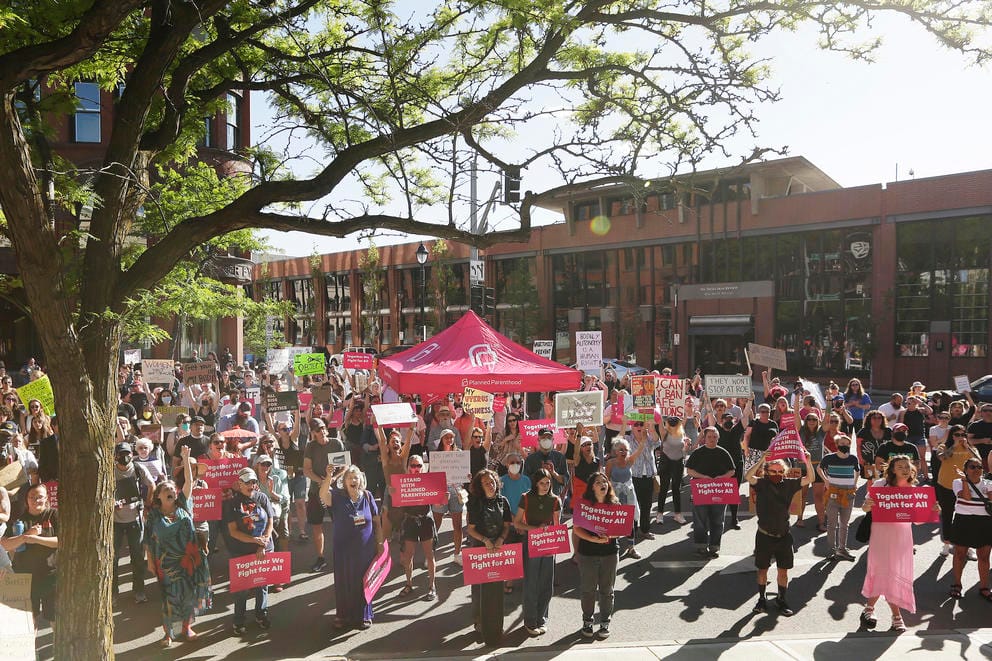 People holding signs with slogans supporting a woman's right to choose an abortion stand in front of a building and a Planned Parenthood tent.
