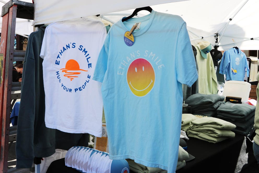 A white shirt hangs next to a blue shirt in a market tent. They both say