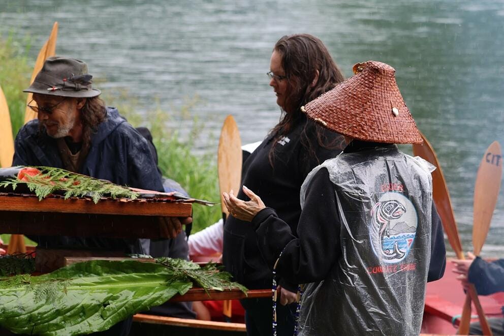 A person wearing a traditional hat and two others stand near a canoe,