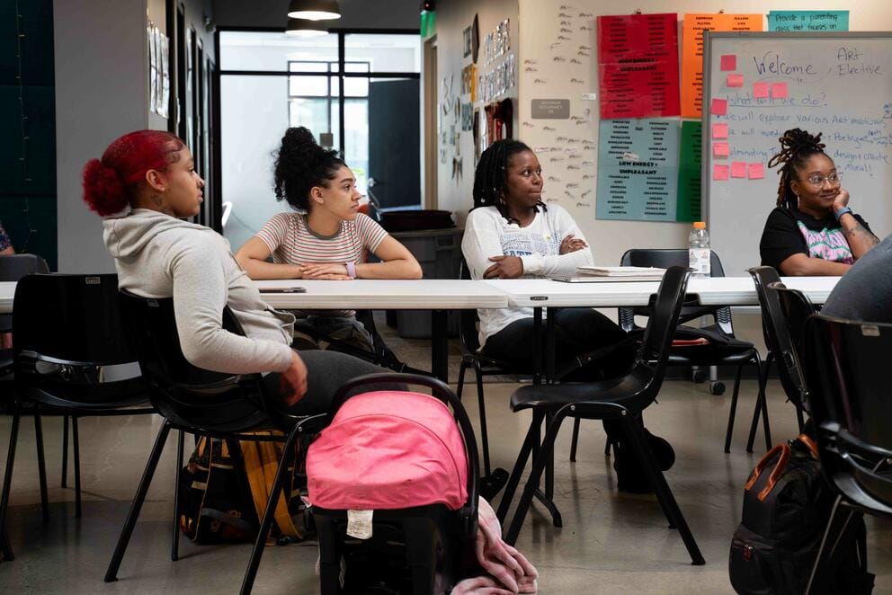 Four high school students pay attention to someone off-screen. There is a baby stroller in the foreground.