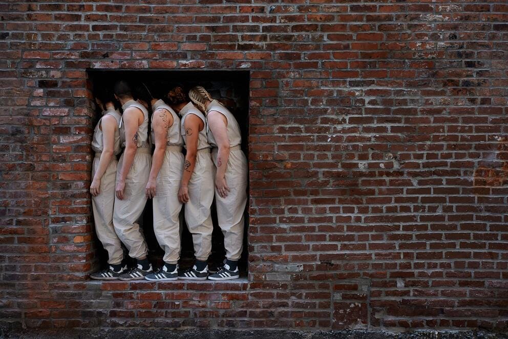 a group of dancers in tan jumpsuits gathers tightly in an opening in a brick wall
