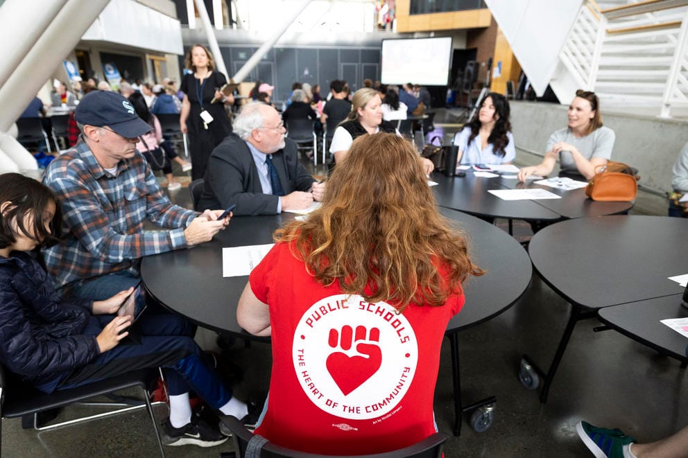 A woman is seen from the back at a table with other people. She is wearing a red shirt that reads