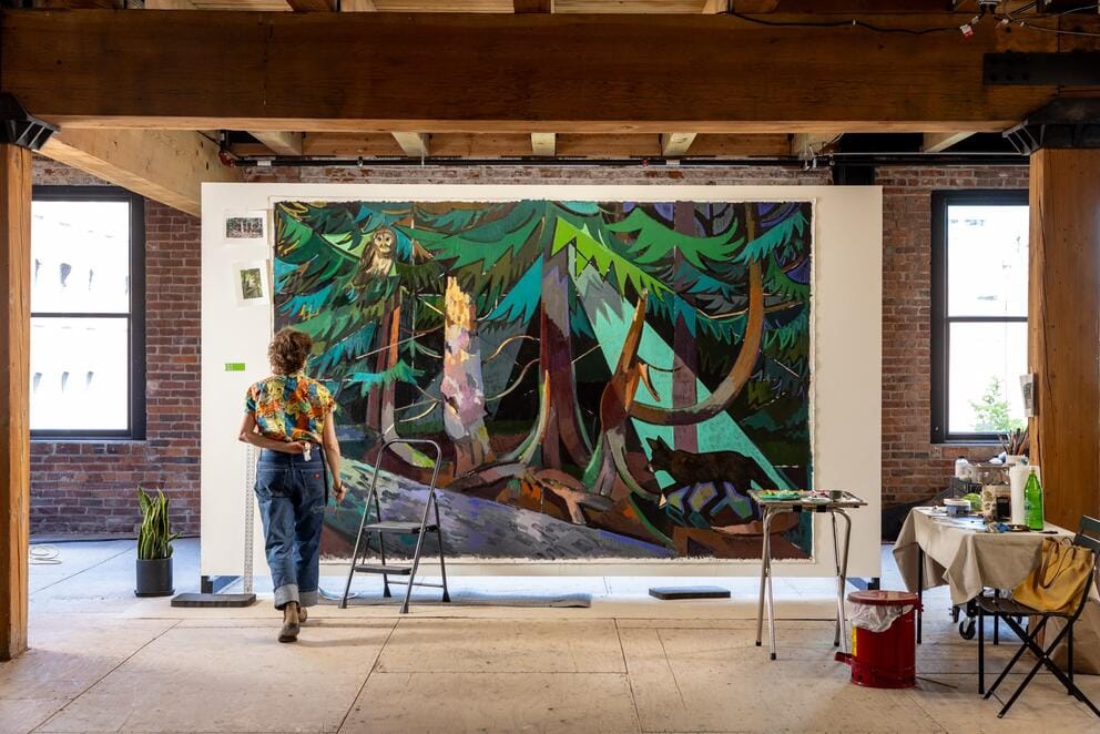 a photo of an artist in an open studio approaching a large painting of a forest