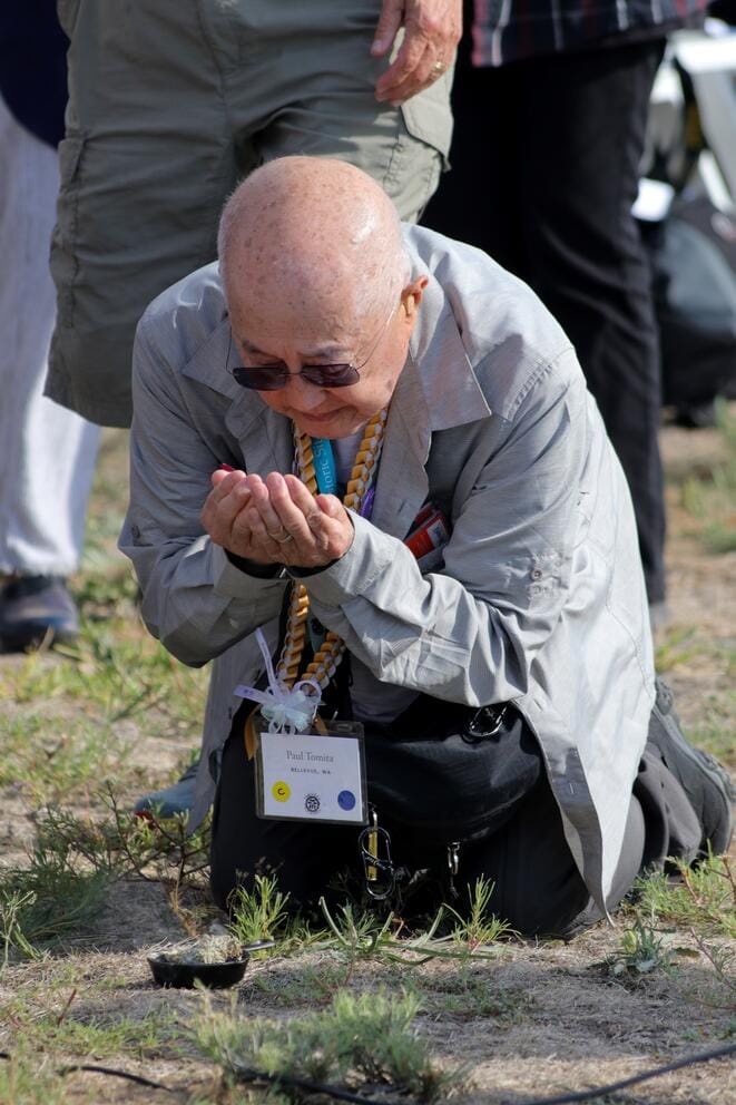 An older person kneels on the ground, palms up in prayer.