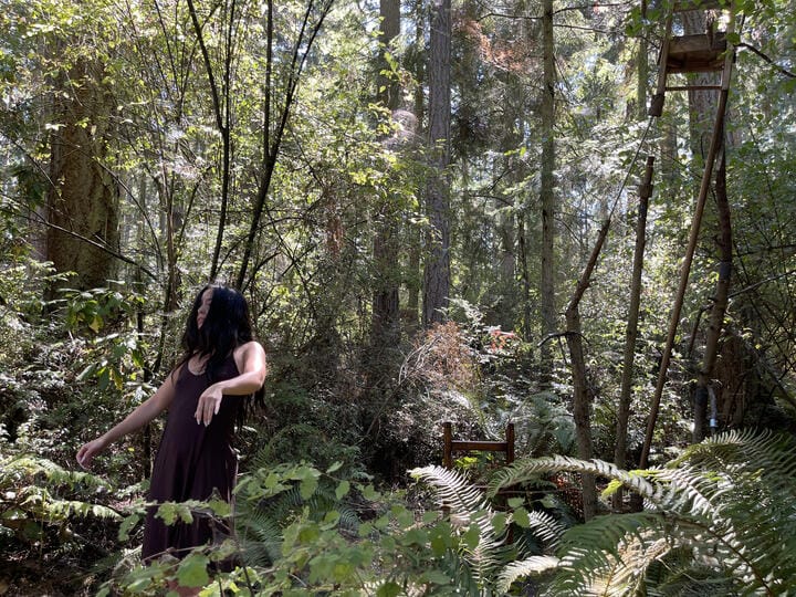 A person in a brown dress with long dark hair stands with their arms slightly akimbo in a sunny, lush forest.