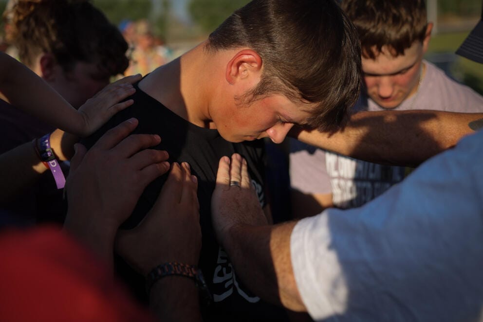 In a tight shot, a young man bows his head as people place their hands on his chest and shoulders