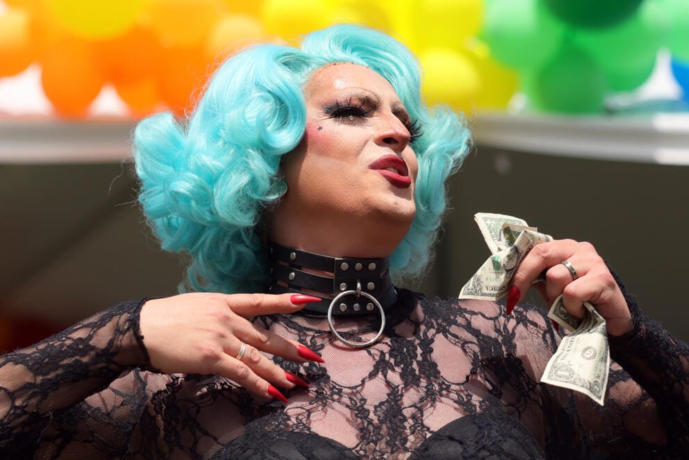 A close up of a drag queen wearing a blue wig and holding a fist full of cash