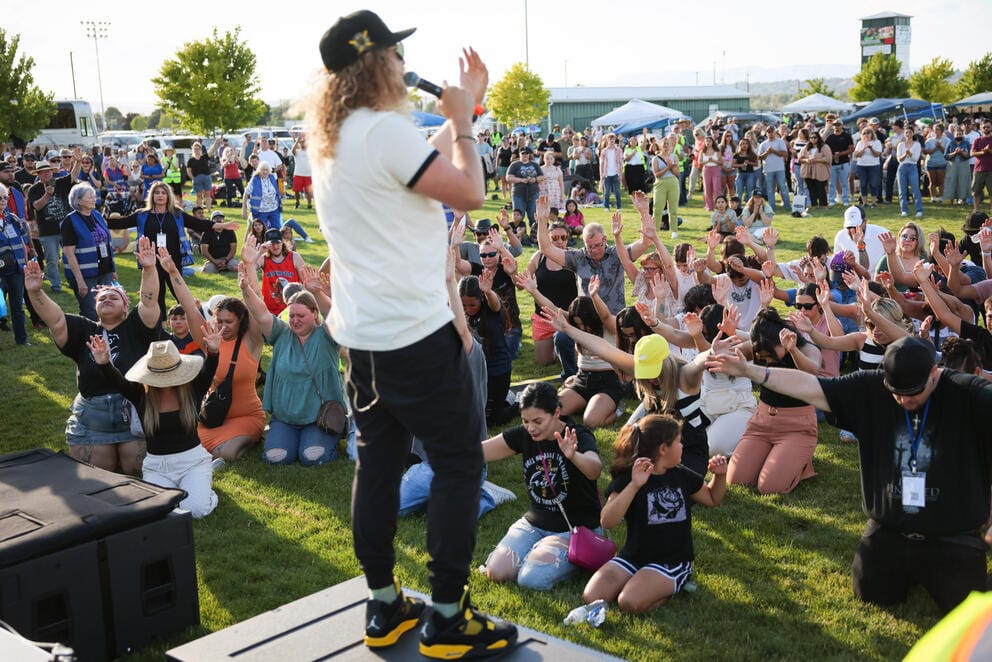 A man stands on stage with a microphone as a crowd of people is on their knees with their hands in the air praying in front of him
