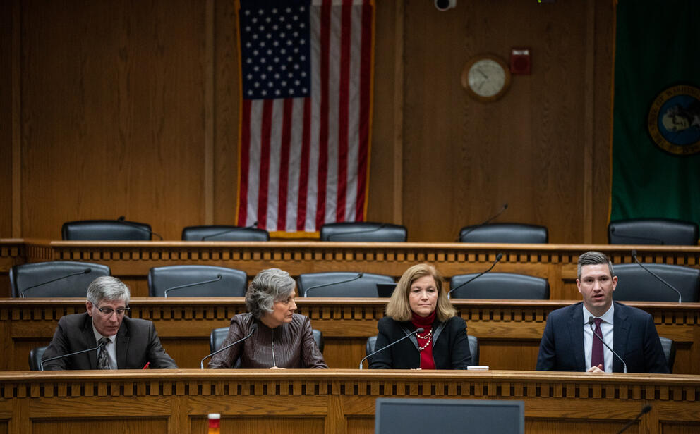 Four people in business attire sitting at a wooden desk in front of an American flag