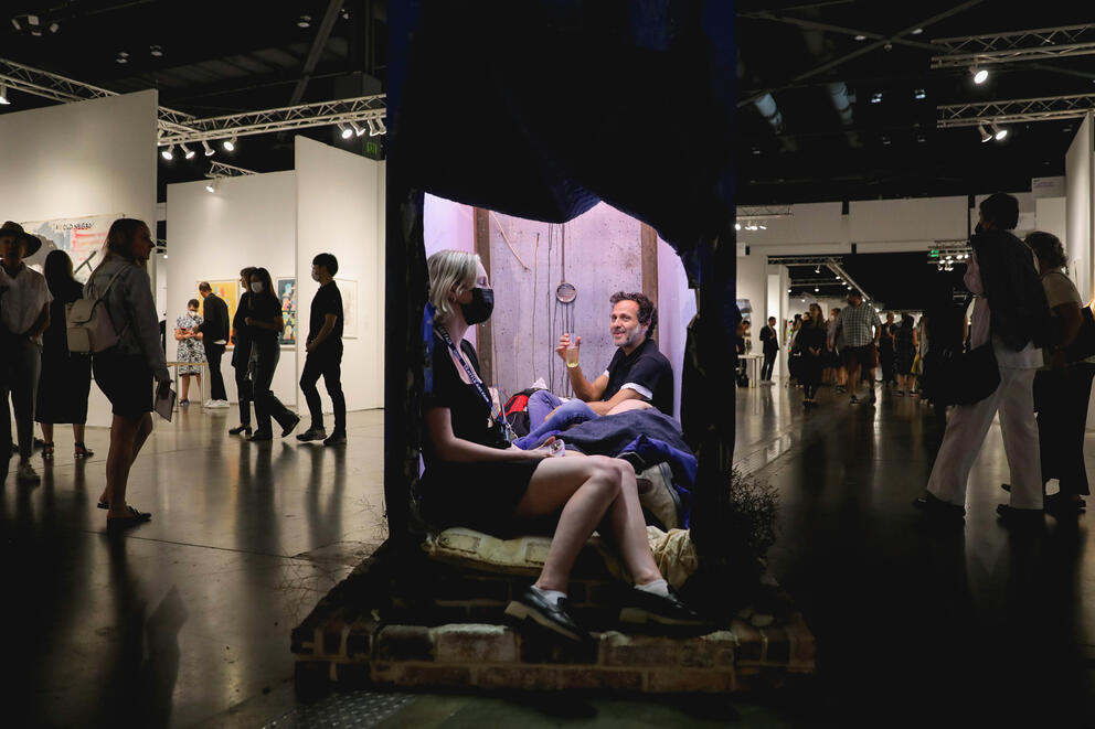 Two people sitting in a lit up small structure while art fair goes on around them