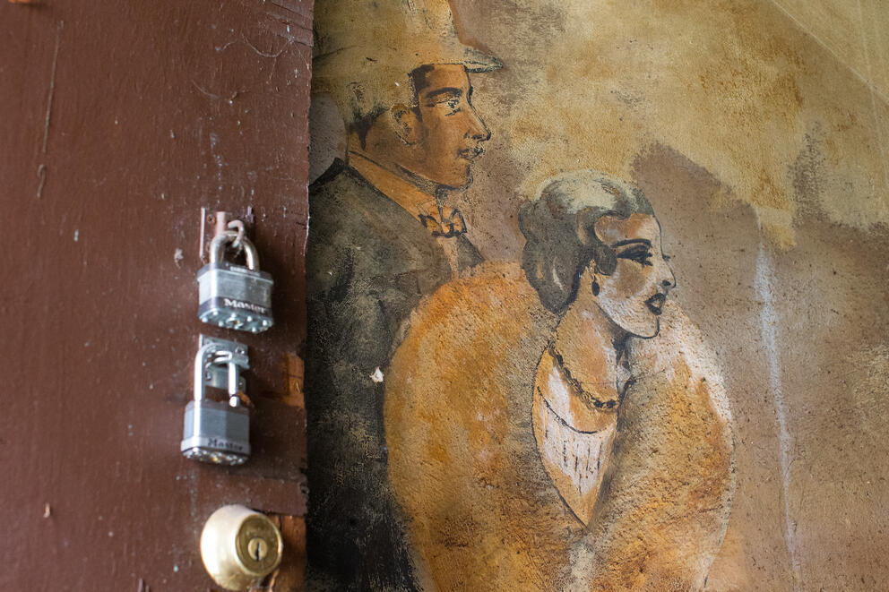 An old mural of a man and a woman is pictured next too a door with padlocks on it.