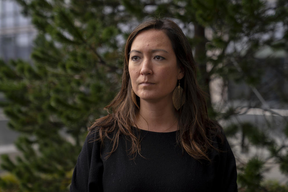 A woman in a black sweater with long brown hair stands in front of trees in background