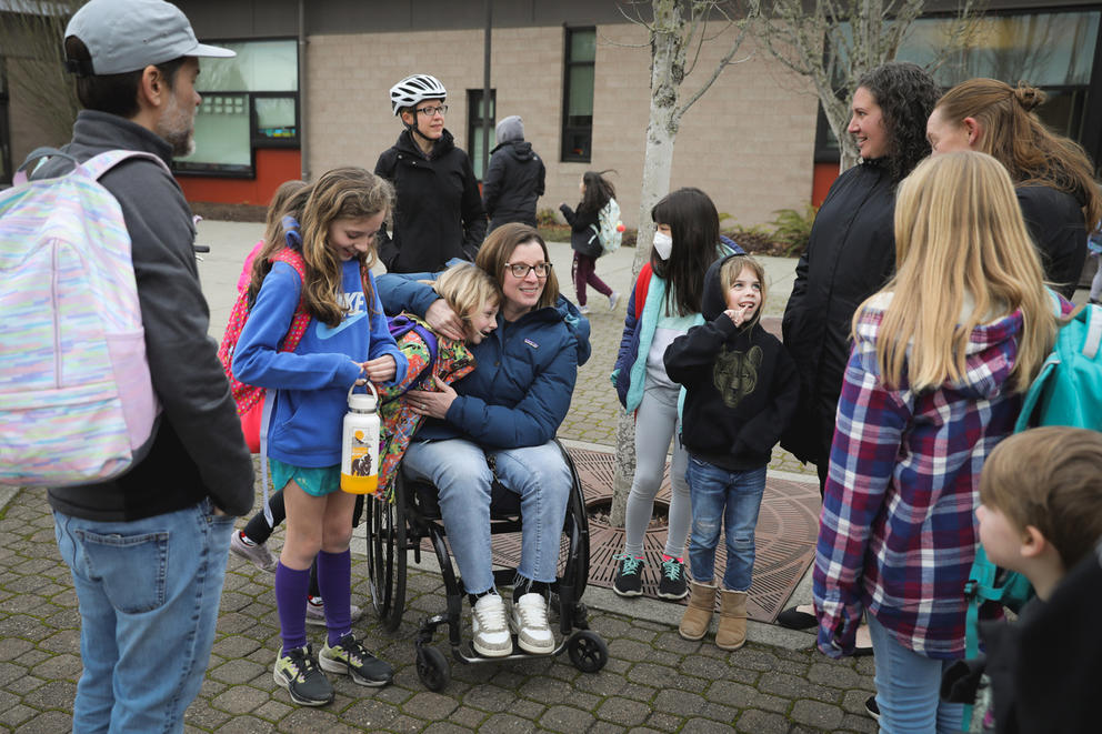 A group of parents and children group together on the sidewalk outside their school.