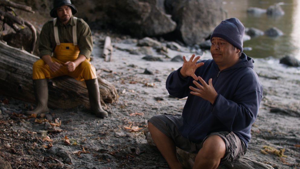 Two people seated on sand, the person in the front is wearing a  hat and gesturing with their hands. The person in teh back to the left is dressed in dungarees and rubber boots, while the person on the right is wearing casual clothes and signing in the foreground as the other person watches. 
