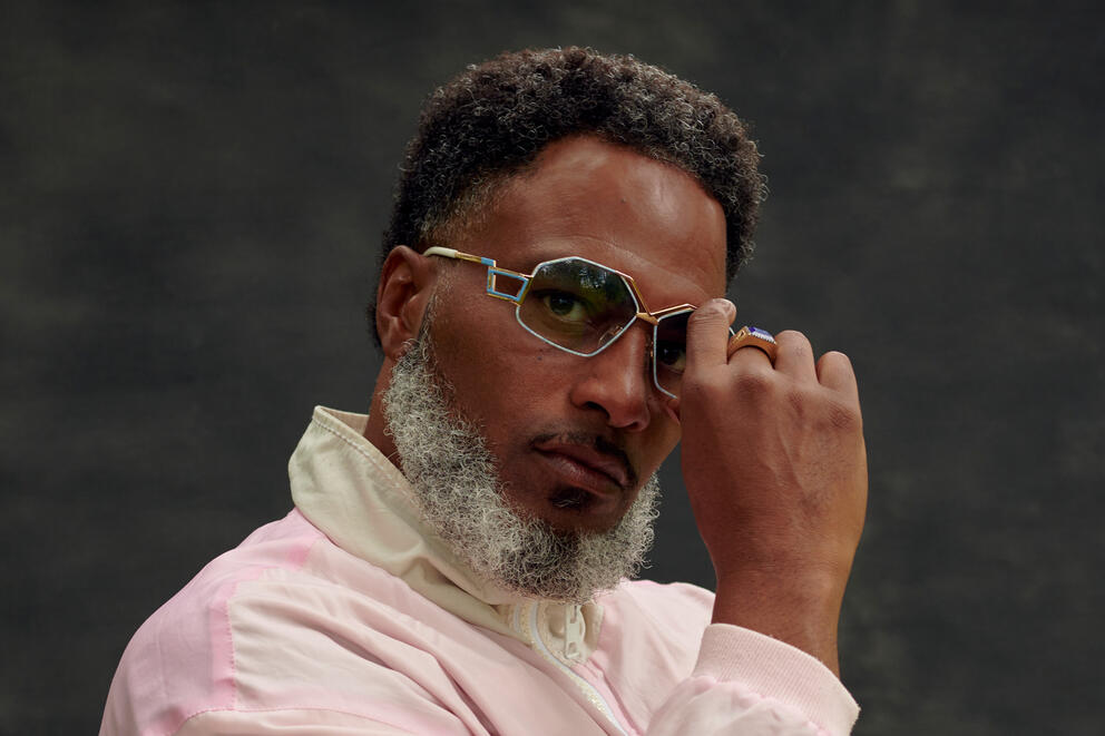 A bearded man looks pensively at the camera as he wears geometric sunglasses