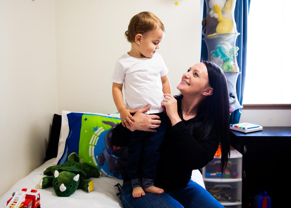 A mother holds her toddler who is standing on a bed