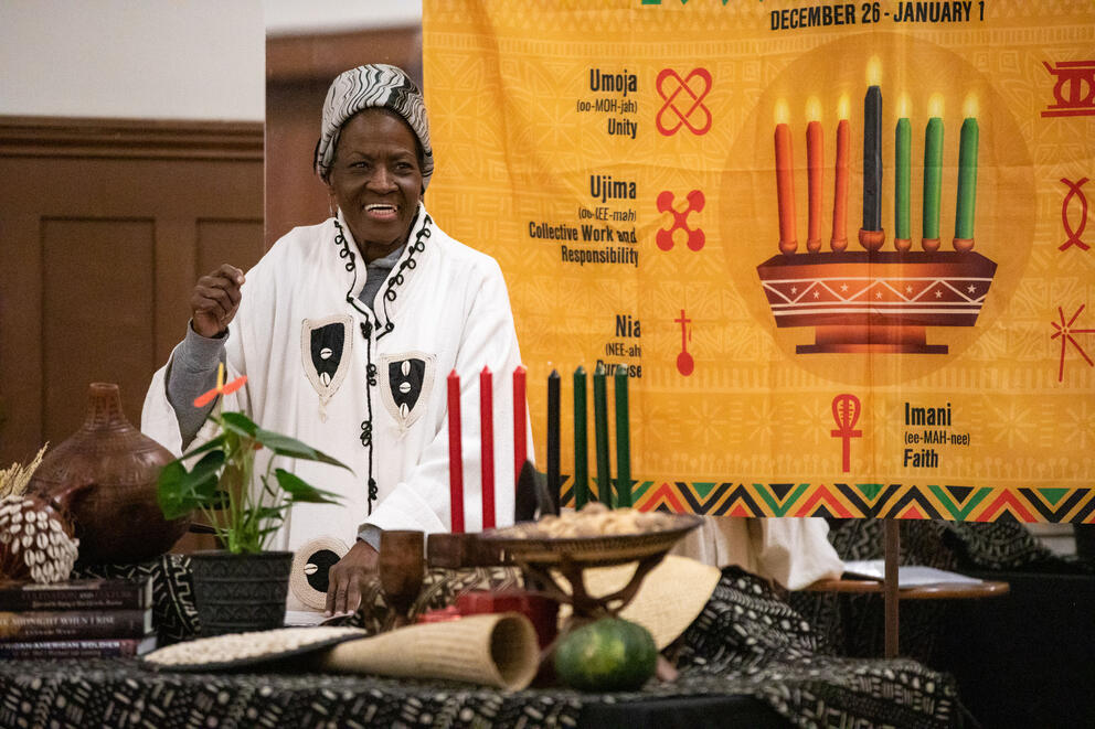 Kbibii Monié dances to African drumming and dancing, behind her hangs a yellow tapestry with candles on it