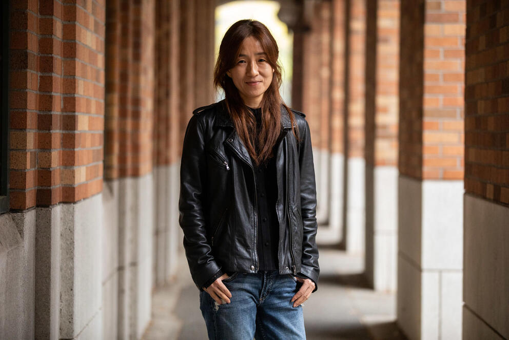 A person wearing a black leather jacket stands between columns and looks at the camera