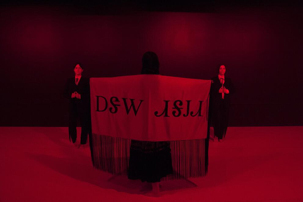 dark stage bathing in red light, one person is wearing with arms wide open a large piece of cloth. Two people in suits stand on either side, a little towards the back.