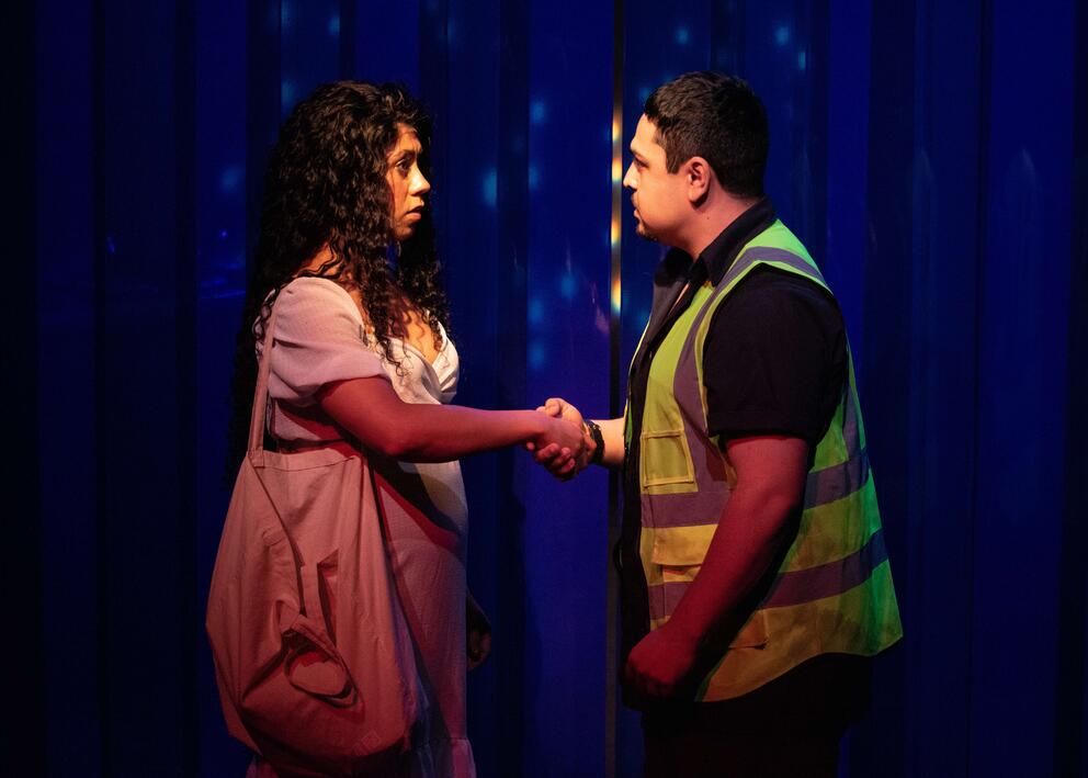 Two people on a stage in front of plastic blue backdrop. She is wearing a white dress and has long, black curly hair. He, shaking her hand, is wearing a yellow hi-viz vest.