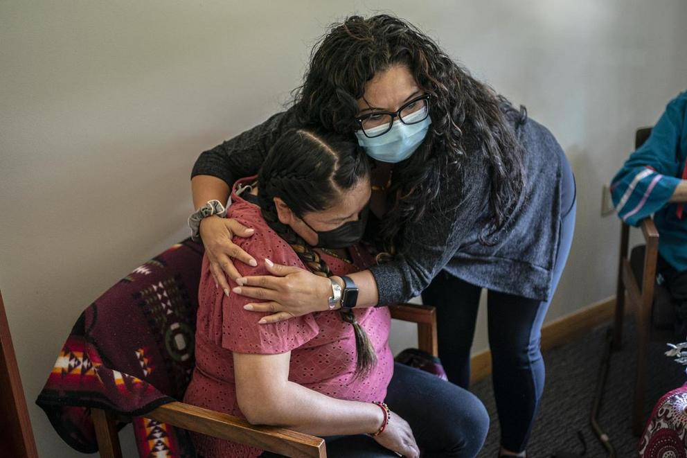 A woman in a face mask gives a seated woman a hug