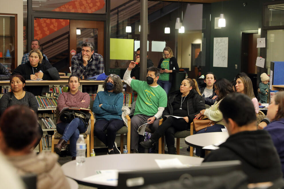 Adults stand and sit at a meeting held in a school library.
