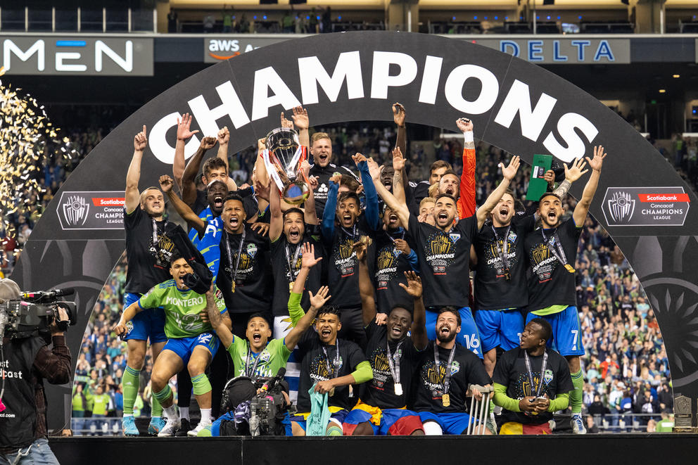 A group of athletes celebrate under a sign reading “CHAMPIONS.”