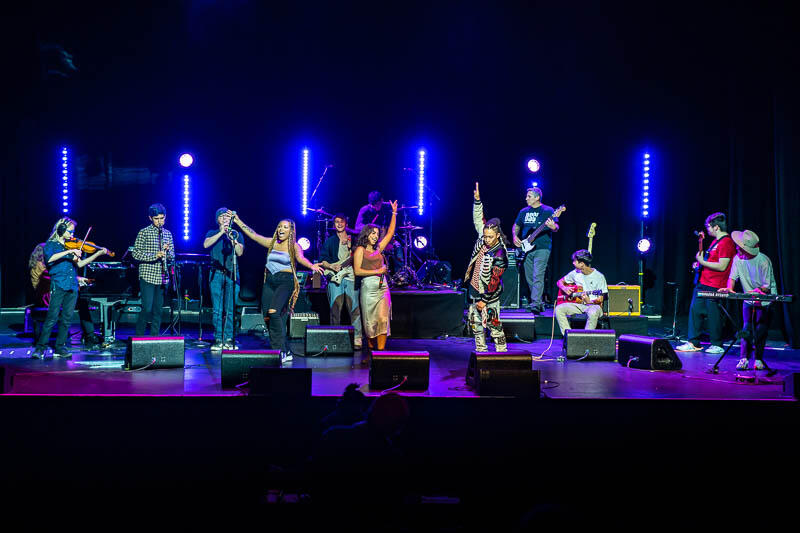 Singers and other musicians stand and perform on stage, blue lights behind them and purple lights on the stage floor