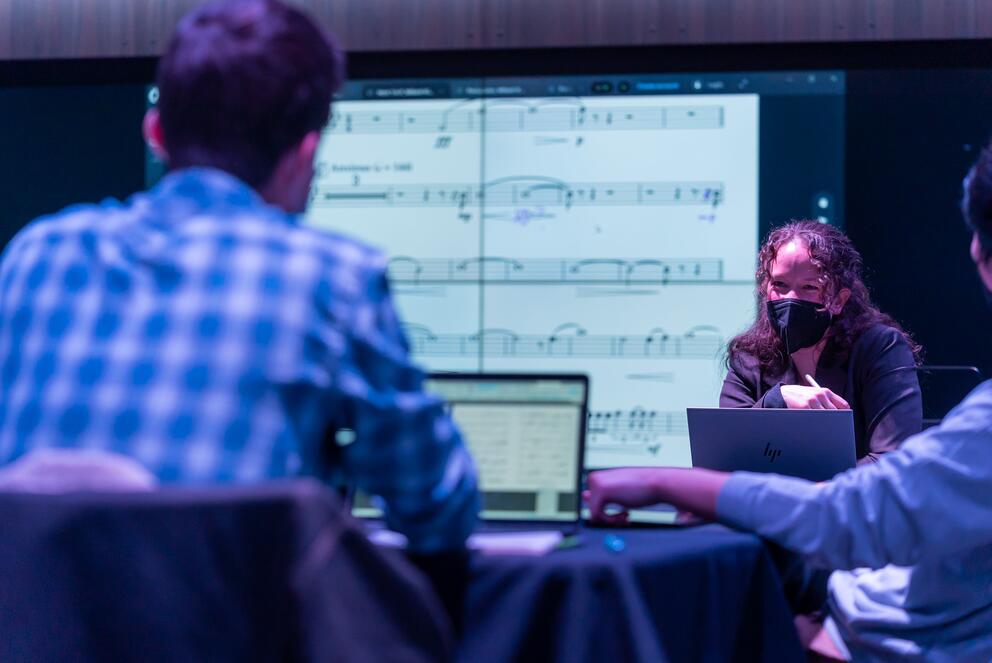 A student sits in front of a teacher. A projection of a score is displayed behind the teacher