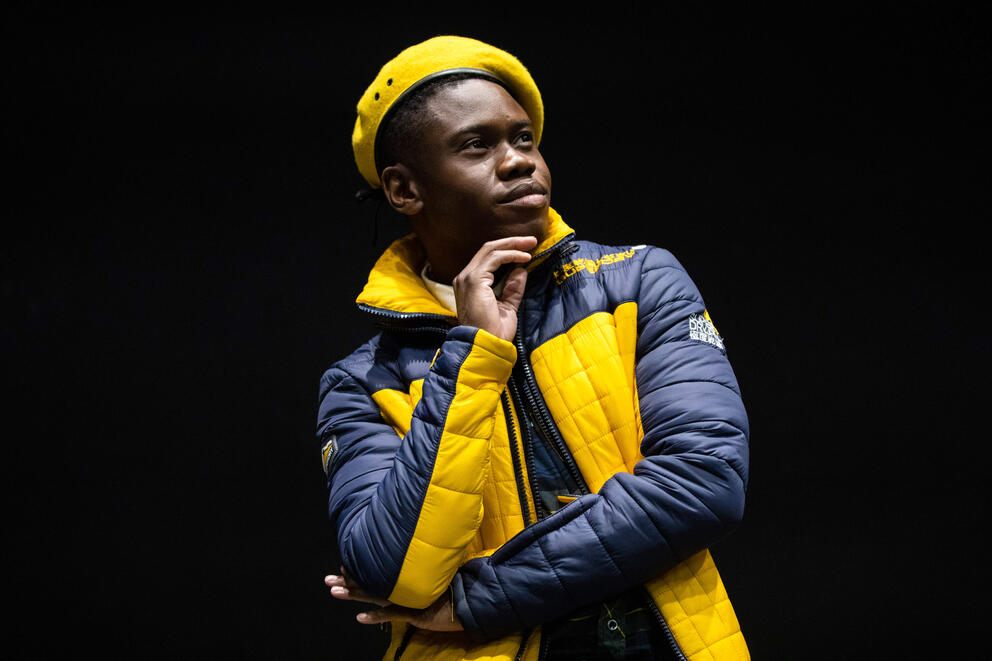 a man in a black and yellow jacket and yellow beret strikes a pose with hand under chin on stage