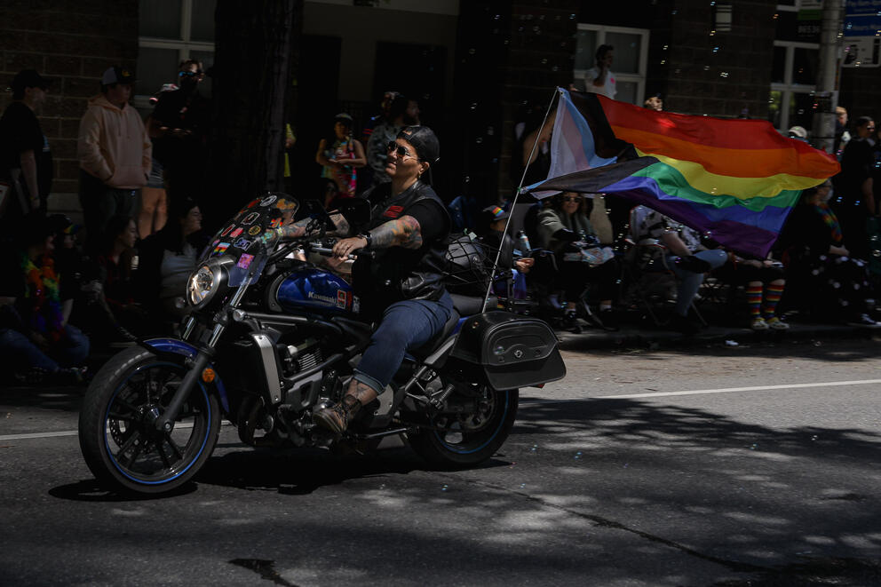 A motorcycle rider is lit up by a patch of sun. A rainbow Pride flag blows behind the bike.