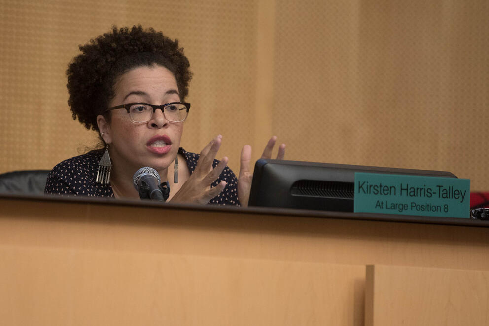 a woman sits behind the city council dais speaking and gesturing with her hands