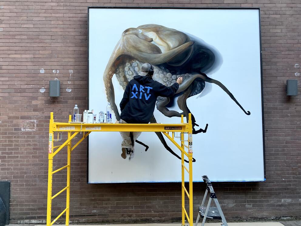 an artist up on a yellow ladder paints an octopus on a white canvas