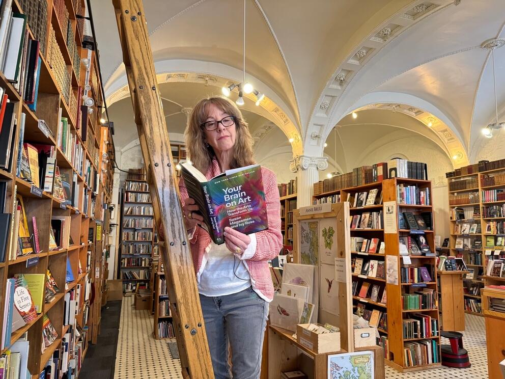 a woman in glasses stands on a ladder reading a book in a book store
