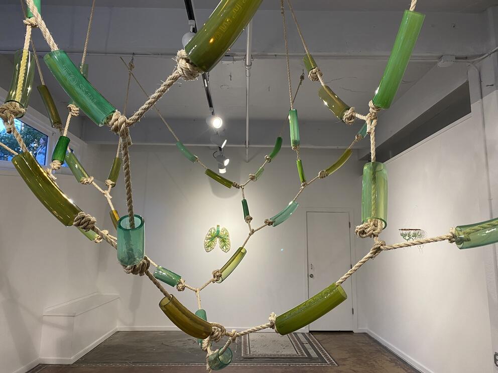 a white walled gallery is hung with hanging ropes partially encased in tubes made of green glass