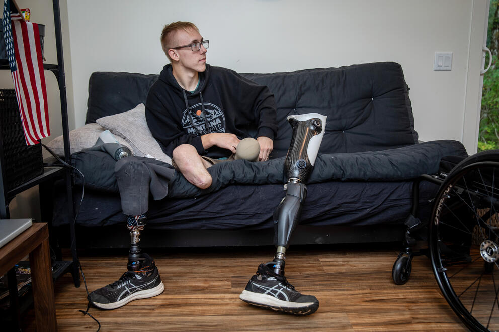 A young man sits on a couch. He is a double amputee and his leg prosthetics are leaned against the couch next to him.