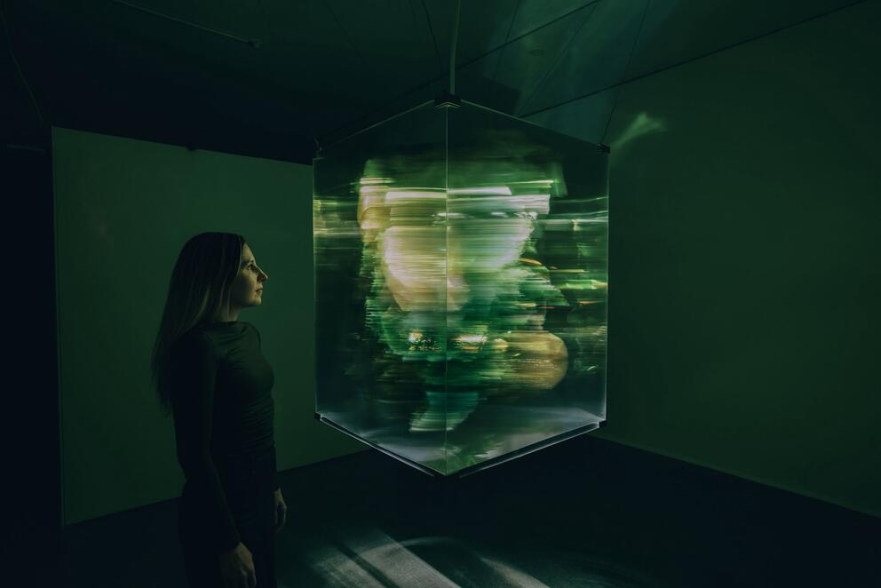 a woman stands in a dark room looking at a floating cube of greenish light