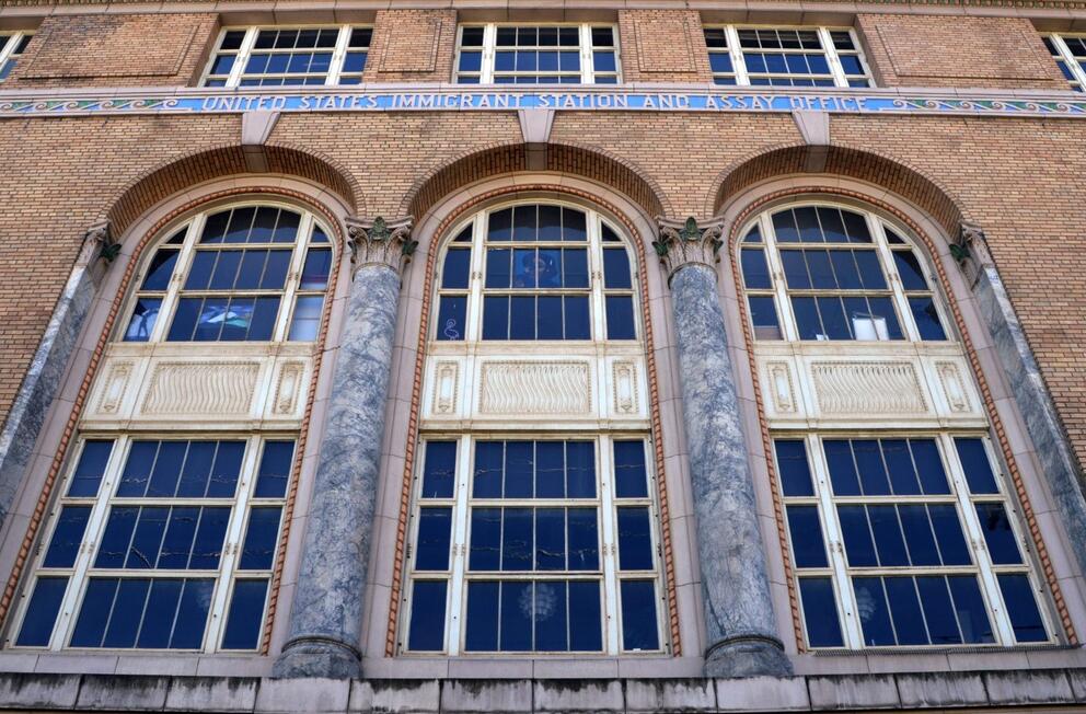 The exterior of the Inscape Arts Building. The building is made from brown bricks with three tall arched windows. "United States Immigrant Station and Assay Office" is written at the top in stone.