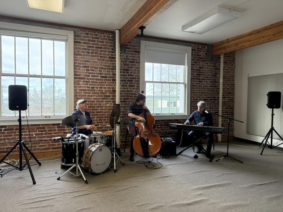 a jazz trio of drums, base and keyboards plays in a brick walled, windowed room