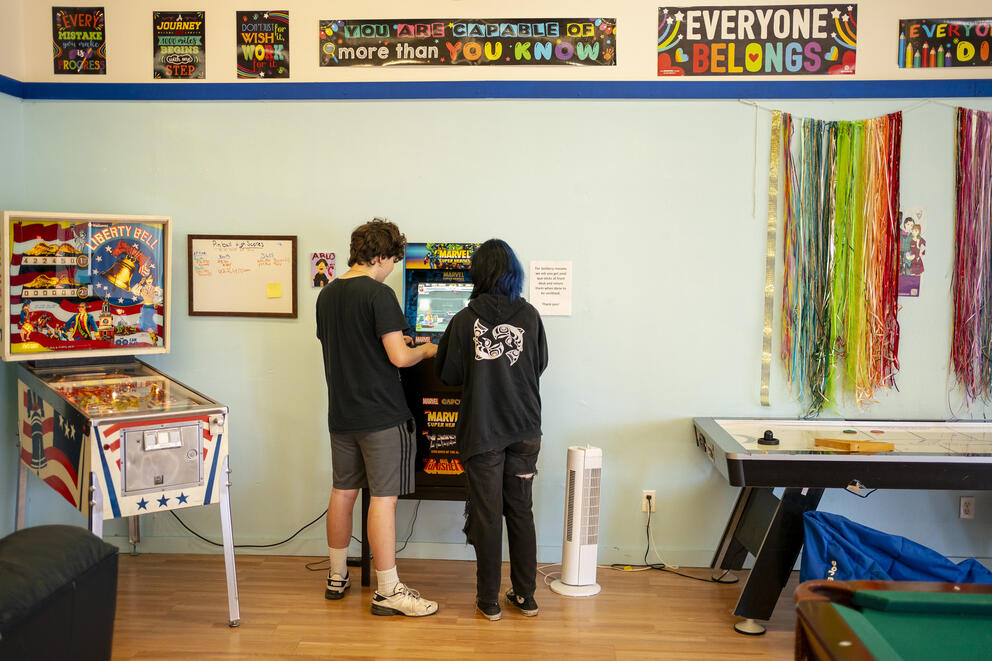Two teens play an arcade game in the corner of a brightly colored youth center