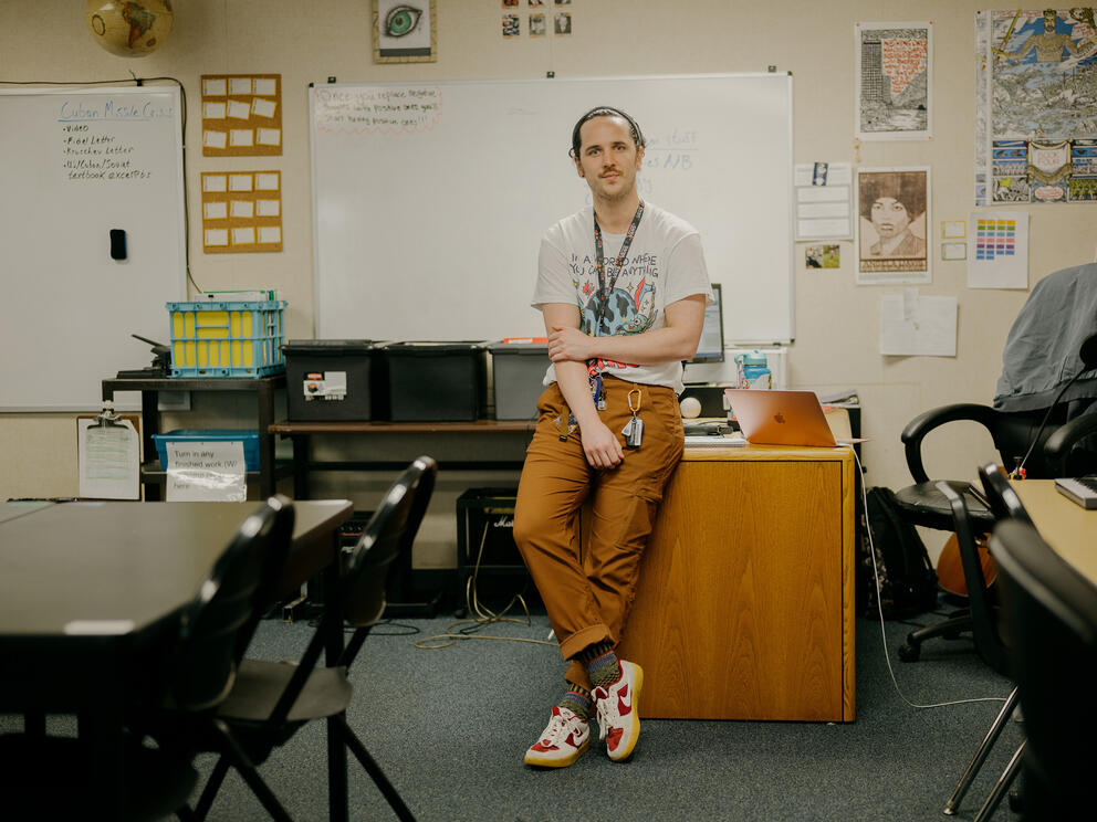 a teacher sits on a desk, looking at the camera