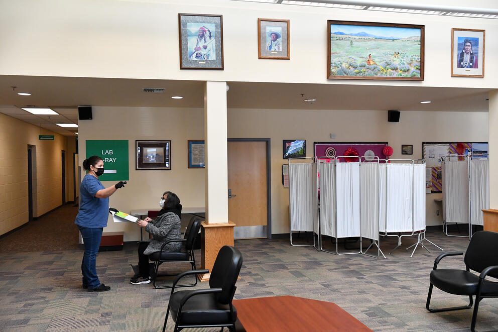A person in scrubs is handing a clipboard to a seated person in a medical clinic type setting.