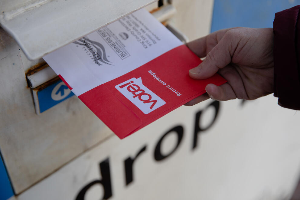 a close up of a person's hand putting a red ballot envelope into a ballot drop box